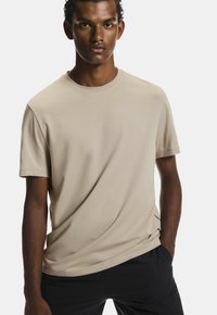 Béžové tričko s okrúhlym výstrihom vyrobené z mäkkého materiálu, s krátkymi rukávmi a voľným strihom, v kombinácii s čiernymi nohavicami.