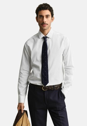 Mann trägt ein weißes Hemd, eine marineblaue Krawatte mit kleinen weißen Punkten, marineblaue Hose und einen geflochtenen braunen Gürtel, hält eine hellbraune Lederjacke.