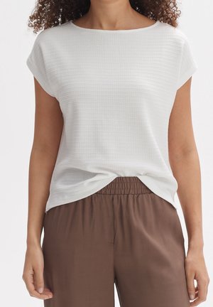 Femme portant un haut blanc texturé à manches courtes légèrement rentré dans un pantalon marron taille élastique sur un fond uni.
