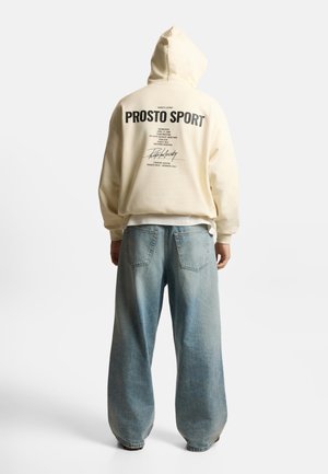 Osoba w kremowej bluzie z kapturem z napisem „PROSTO SPORT” na plecach i luźnych jasnoniebieskich dżinsach, stojąca tyłem na białym tle.