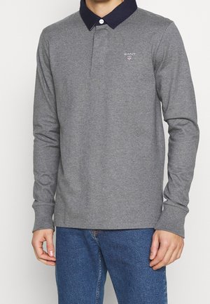 Mężczyzna ubrany w szarą, długorękawową koszulkę polo z ciemnogranatowym kołnierzem i niebieskie dżinsy, stojący na jednolitym tle.