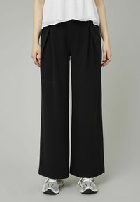 Pantalons larges noirs en tissu lisse, dotés de plis à la taille. Associés avec des baskets grises pour un look décontracté.