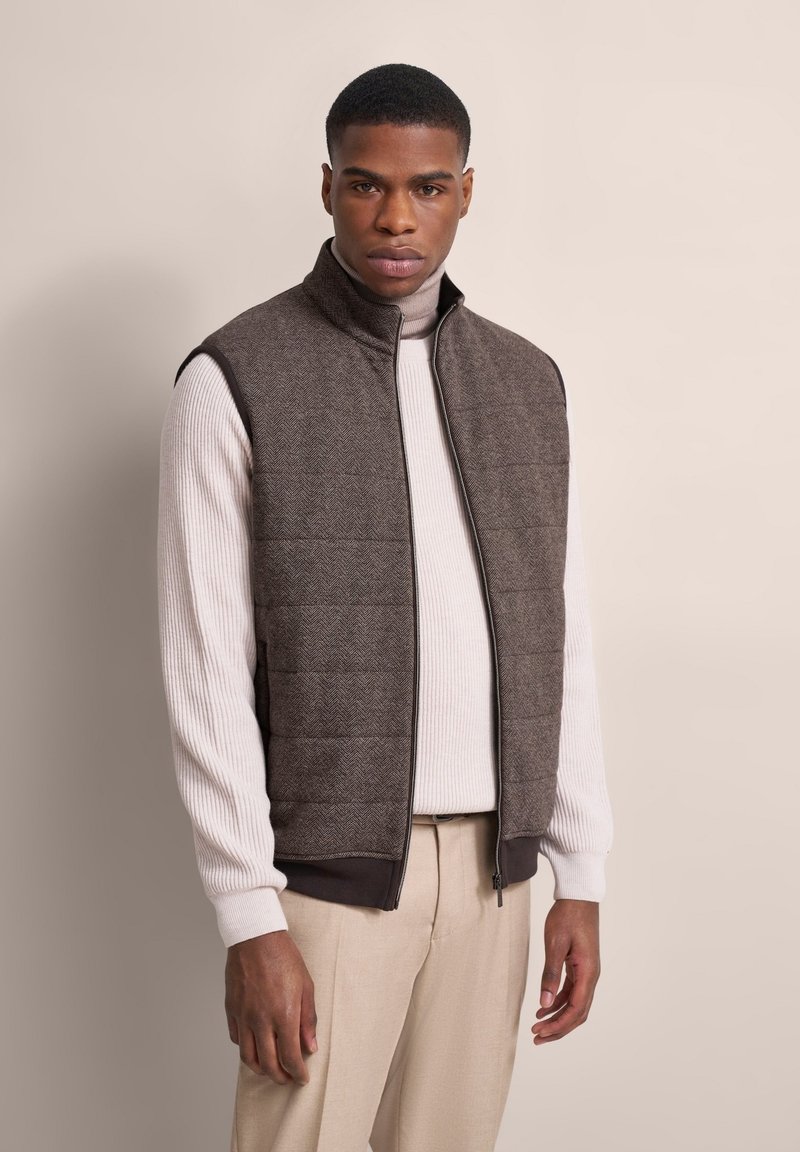 Braune Fischgrät-Muster ärmellose Weste mit hohem Kragen, Reißverschluss und gerippten Kanten, getragen über einem cremefarbenen Rollkragenpullover und beigen Hosen.