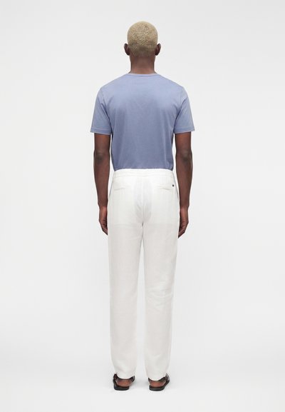 Homme aux cheveux blonds courts, debout de dos, portant une chemise à manches courtes bleu clair, un pantalon blanc et des sandales foncées sur un fond uni.