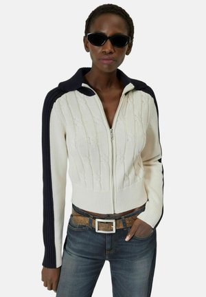 Kvinde iført sorte katteøje-solbriller, creme- og sort striktrøje med lynlås, brun bælte og mørkeblå jeans, poserer med den ene hånd i lommen.