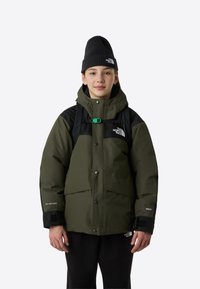 Giacca isolante verde con cappuccio e dettagli neri, dotata di cerniera frontale e bottoni a pressione. Completa il look un berretto nero.