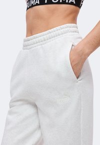 Κοντινό πλάνο ατόμου που φοράει λευκή φόρμα Puma με το χέρι στην τσέπη και μαύρο αθλητικό σουτιέν Puma.