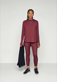 Bordeaux idrottshoodie med ficka fram, matchad med texturerade leggings med ett mönster. Svarta sneakers och en handduk i handen.