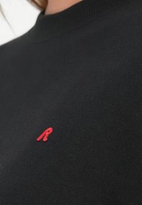 Svart hettegenser med høy rund hals og en liten rød brodert "R"-logo på venstre brystområde. Myk stofftekstur.