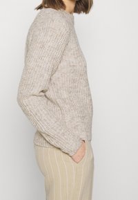Femme portant un pull en maille côtelée beige à manches longues, associé à un pantalon beige avec des rayures verticales blanches, vue de profil avec la main dans la poche.