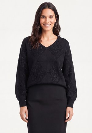 Kvinde med mørkt, bølget hår bærer store øreringe i ringform, en sort V-hals sweater med glimmerdetaljer og et sort nederdel, smilende til kameraet.