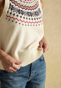 Adīta krēmkrāsas bezšķiedras topa ar rakstainu yoke, kurā ir sarkanie, zilie un brūnie akcenti. Ribots apakšmalas un brīva piegriezuma kombinācija ar zilu denim džinsu.