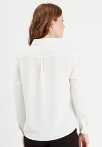 Chemise blanche à manches longues en tissu texturé, col avec un détail de pli au dos. Matériau lisse avec ourlet arrondi.