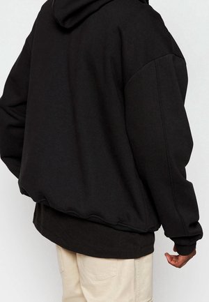 Personne portant un sweat à capuche noir ample par-dessus un t-shirt noir associé à un pantalon beige clair, vue de dos et de côté gauche.
