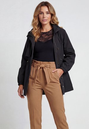 Vrouw met golvend blond haar, gekleed in een zwart jasje over een zwart kanten top en hoge taille beige broek met een gestrikte riem, staand met één hand in haar zak.