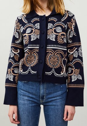 Femme portant un cardigan bleu marine boutonné avec des motifs floraux beige et blancs et un jean bleu, raccourci aux hanches.