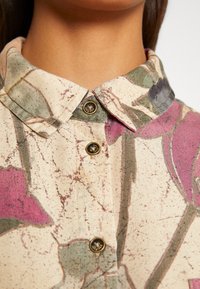 Gros plan d'une chemise à col avec un motif floral abstrait beige, rose et vert et trois boutons en laiton visibles à l'avant.
