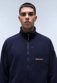Marineblaue Fleecejacke mit hohem Kragen, Reißverschlusstasche und markenbezogenen Akzenten. Die weiche Textur und das athletische Design betonen die Funktionalität.
