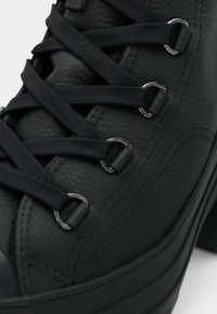 Gros plan d'une chaussure en cuir noir avec des lacets noirs passés dans des œillets métalliques et des détails cousus sur le côté.
