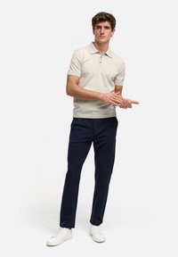 Beige polobotka s krátkým rukávem a třemi knoflíky, nošena s tmavě modrými kalhotami a bílými teniskami. Model stojí s odkrytýma rukama.