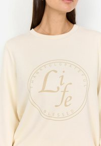 Vrouw in een crème sweatshirt met een ronde beige afdruk met de tekst "Life" en de woorden "Thankful", "Grateful" en "Blessed" eromheen.