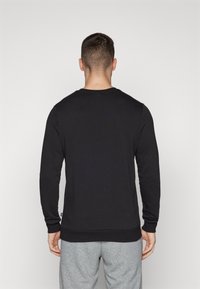 Hombre de pie de espaldas, vistiendo una camisa negra de manga larga y pantalones grises, contra un fondo claro y liso.