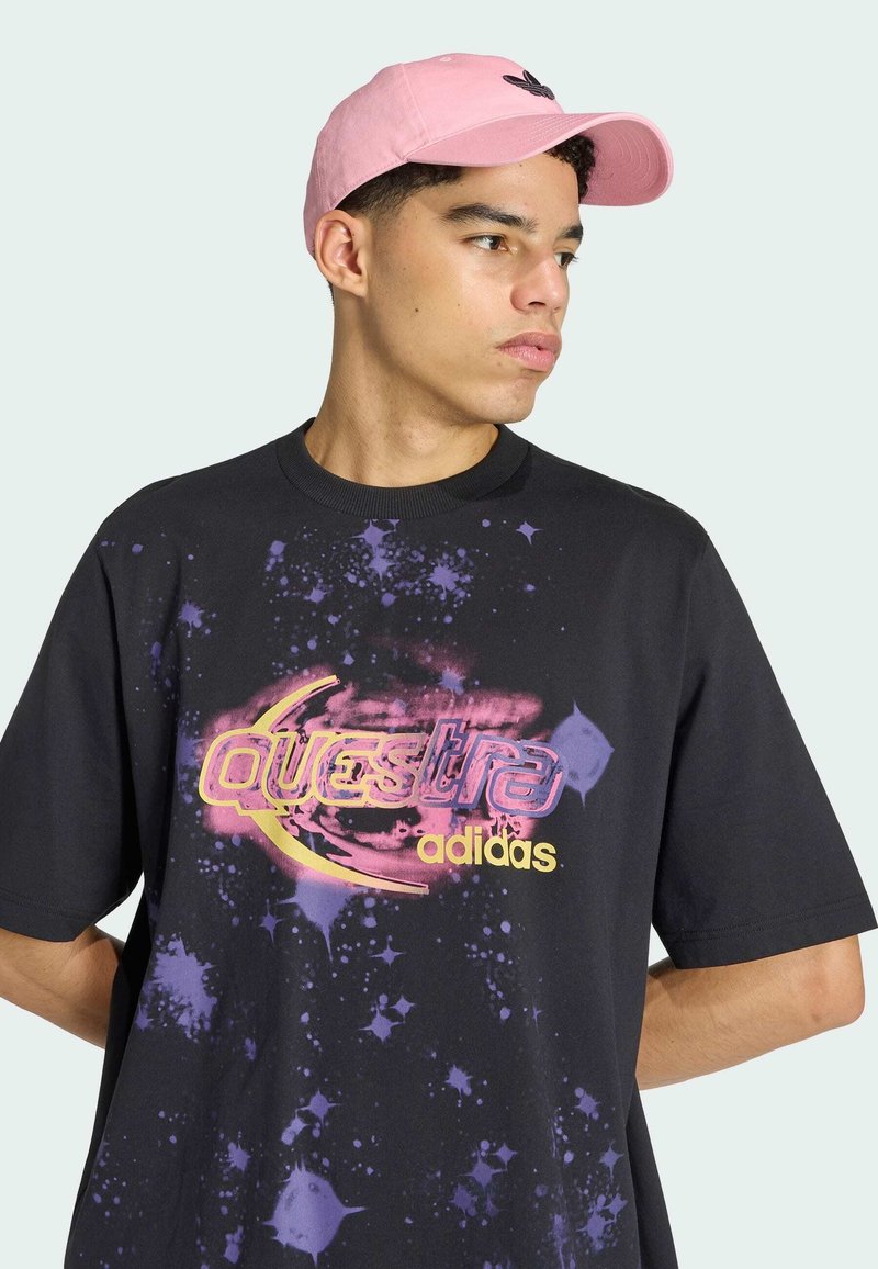 Junger Mann mit rosa Kappe und schwarzem Adidas-Shirt mit lila Galaxie-Print und der Aufschrift „Questra“, schaut nach rechts vor einfachem Hintergrund.