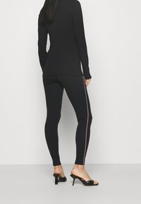 Zwarte lange mouw top en aansluitende legging met een zijstripe in het rood, gecombineerd met zwarte open teen hoge hakken. Glad materiaal, smalle pasvorm.