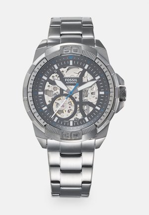 Ceas automat Fossil cu brățară din oțel inoxidabil argintiu, cadran negru, display scheletat, accente albastre și bezel înclinat cu marcaje clare.