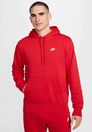 Hombre con sudadera y pantalones deportivos rojos a juego de Nike con logotipos blancos, de pie con una mano en el bolsillo delante de un fondo liso.