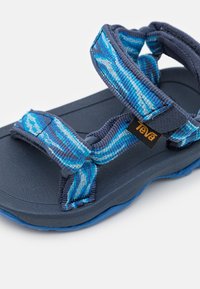 Teva-sandal med en mørkeblå gummisåle, blå mønstrede stoffremmer og en justerbar lukking. Merkbar logo på remmen.