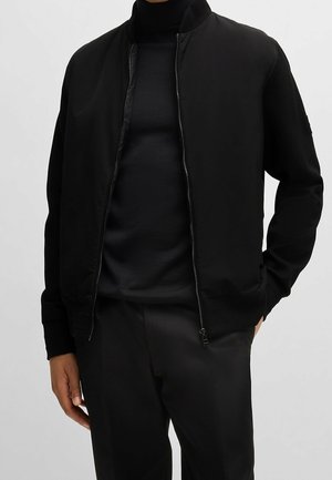 Personne portant une veste bomber noire zippée sur un col roulé noir et un pantalon noir, debout, une main dans la poche.