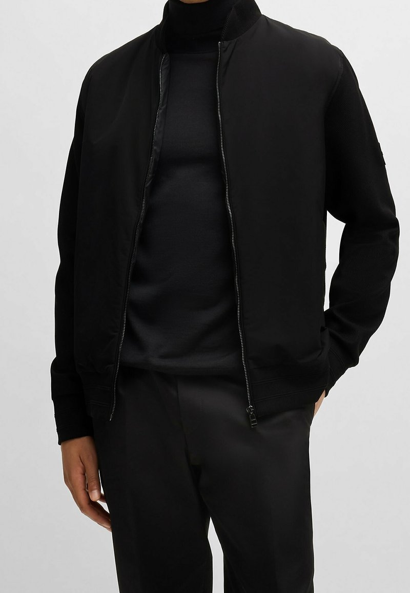 Personne portant une veste bomber noire zippée sur un col roulé noir et un pantalon noir, debout, une main dans la poche.