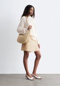 Femme aux cheveux bouclés portant un pull crème, une jupe beige, des chaussures blanches à bouts ouverts et bride arrière, tenant un sac à bandoulière beige, debout à l'intérieur.