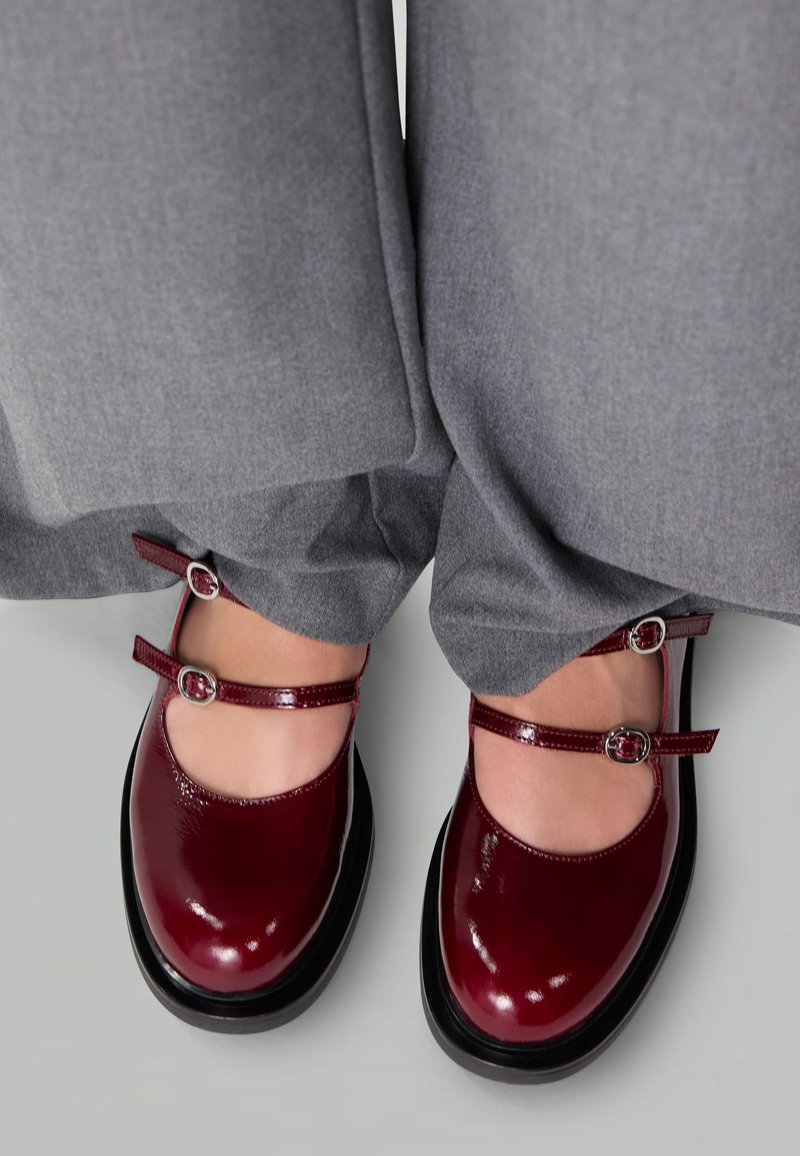 Glänzende burgunderfarbene Mary Jane-Schuhe mit doppelten Riemen und silbernen Schnallen, getragen mit lockeren grauen Hosen auf einem hellgrauen Boden.