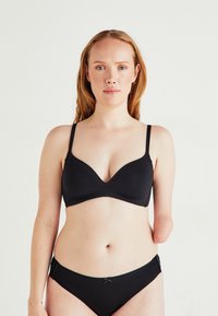 Woman with light skin and red hair wearing black bra and underwear, facing forward, with left arm below elbow missing, plain white background.