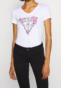 Camiseta blanca de manga corta con cuello en V, que presenta un gráfico floral y geométrico en rosa y verde con el texto "GUESS U.S.A." en el centro.