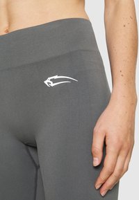 Szare legginsy wykonane z elastycznego materiału, z wysokim stanem i białym akcentem logo na biodrze. Gładka faktura z płaskimi szwami.