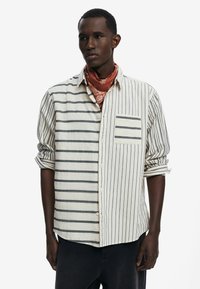Camisa de manga larga a rayas en crema claro con rayas oscuras, que presenta un bolsillo en el pecho y las mangas arremangadas. Material de algodón.
