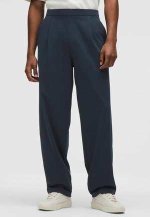 Person iført løstsiddende mørkeblå bukser og hvide sneakers, stående mod en ensartet lys baggrund.