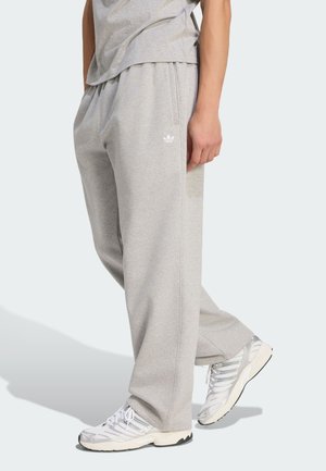 Person, die hellgraue Adidas-Sweatpants und weiße Sportschuhe trägt, steht mit einem leicht angewinkelten Bein.
