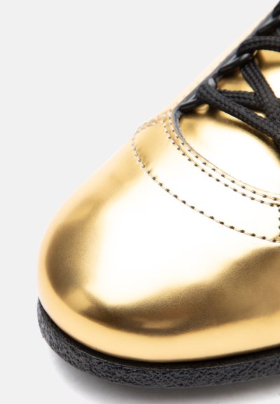 Detailný záber zlatej kovovej špičky topánky s čiernou textúrovanou podrážkou a čiernymi šnúrkami na bielom pozadí.
