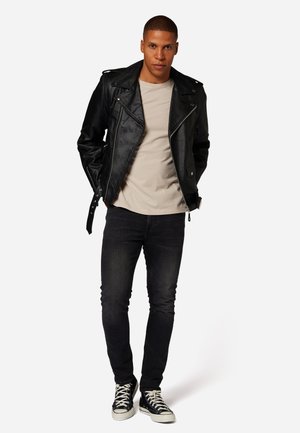 Veste de moto en cuir noir avec des éléments en métal argenté, t-shirt beige, jeans foncés et baskets noires, pose debout devant un fond uni.