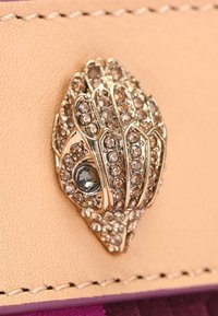 Gouden en rhinestone slangenkopdetail op tan leer met zichtbare stiksels en een paarse rand eronder.