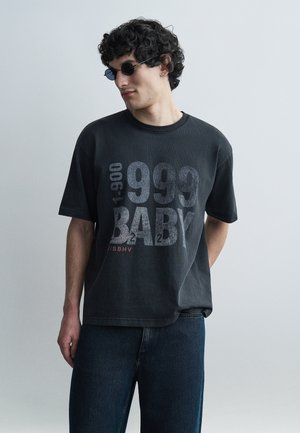 Νεαρός άντρας με σγουρά μαλλιά που φοράει στρογγυλά μαύρα γυαλιά ηλίου, μια ξεθωριασμένη μαύρη υπερμεγέθη μπλούζα με το κείμενο "1999 BABY" και σκούρα τζιν.