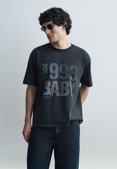 Joven con cabello rizado que lleva gafas de sol redondas negras, una camiseta negra desgastada de gran tamaño con el texto "1999 BABY" y jeans oscuros.