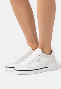 Baskets blanches en matériaux daim et cuir, à bout rond, avec un laçage et une bande noire sur la semelle, arborant un logo sur le côté.