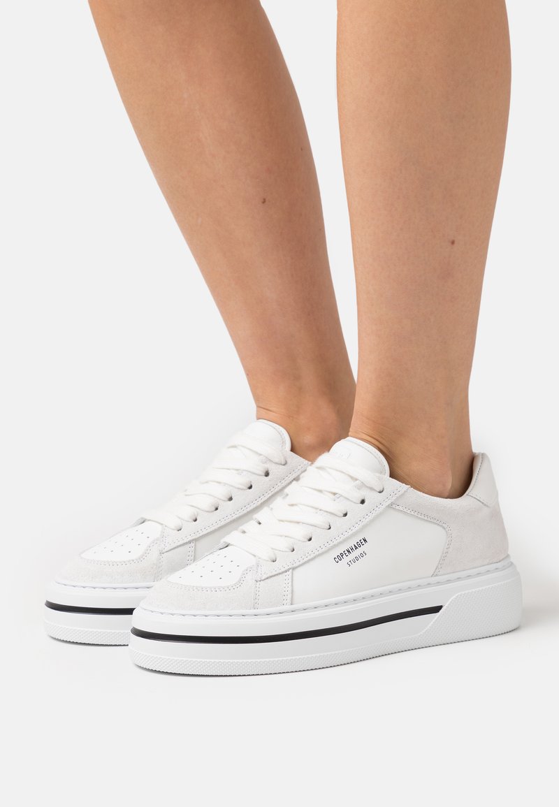 Baskets blanches en matériaux daim et cuir, à bout rond, avec un laçage et une bande noire sur la semelle, arborant un logo sur le côté.