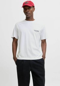 White cotton t-shirt with short sleeves, featuring a printed design on the left chest. Paired with black trousers, model wears a red cap.