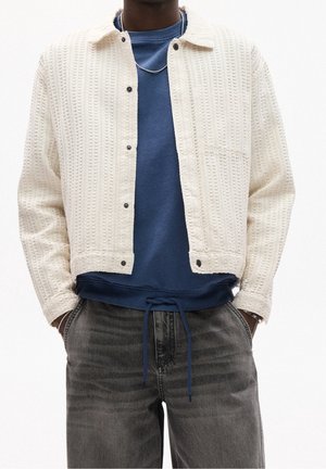 Persona lleva una chaqueta cremosa con textura y botones sobre una sudadera azul con cordones y vaqueros grises, manos en los bolsillos.